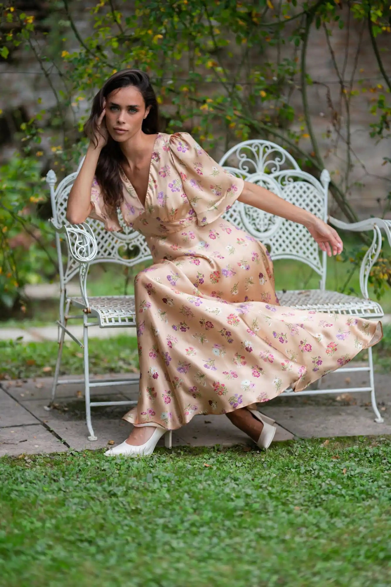 A woman in a flowing, floral print maxi dress and white high heels, seated on a white ornate bench.