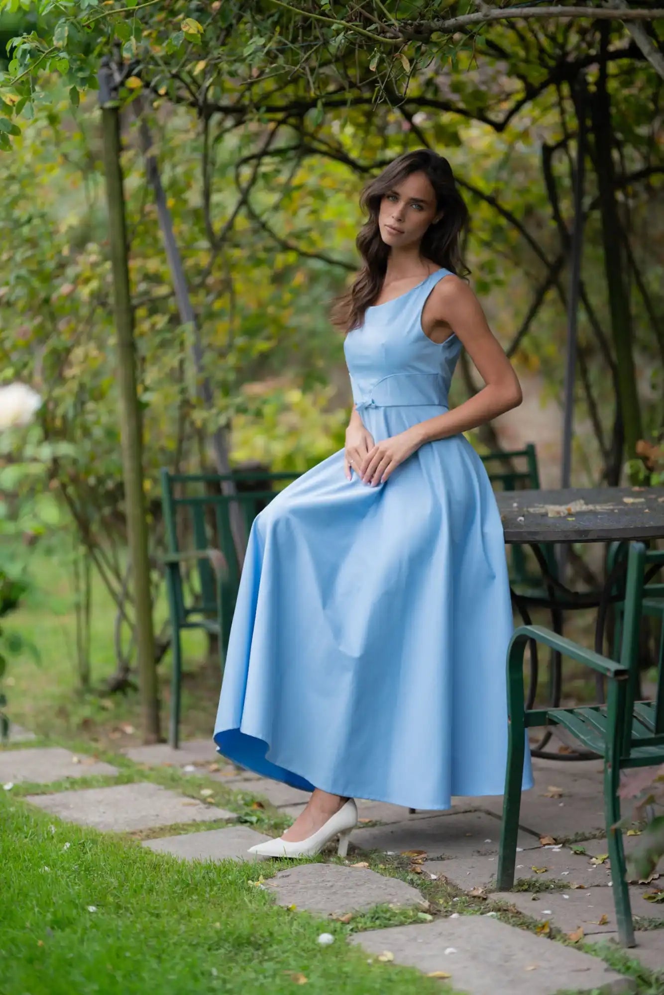 A woman in a light blue sleeveless maxi dress with a fitted bodice and a full, flowing skirt, paired with white pointed-toe heels.