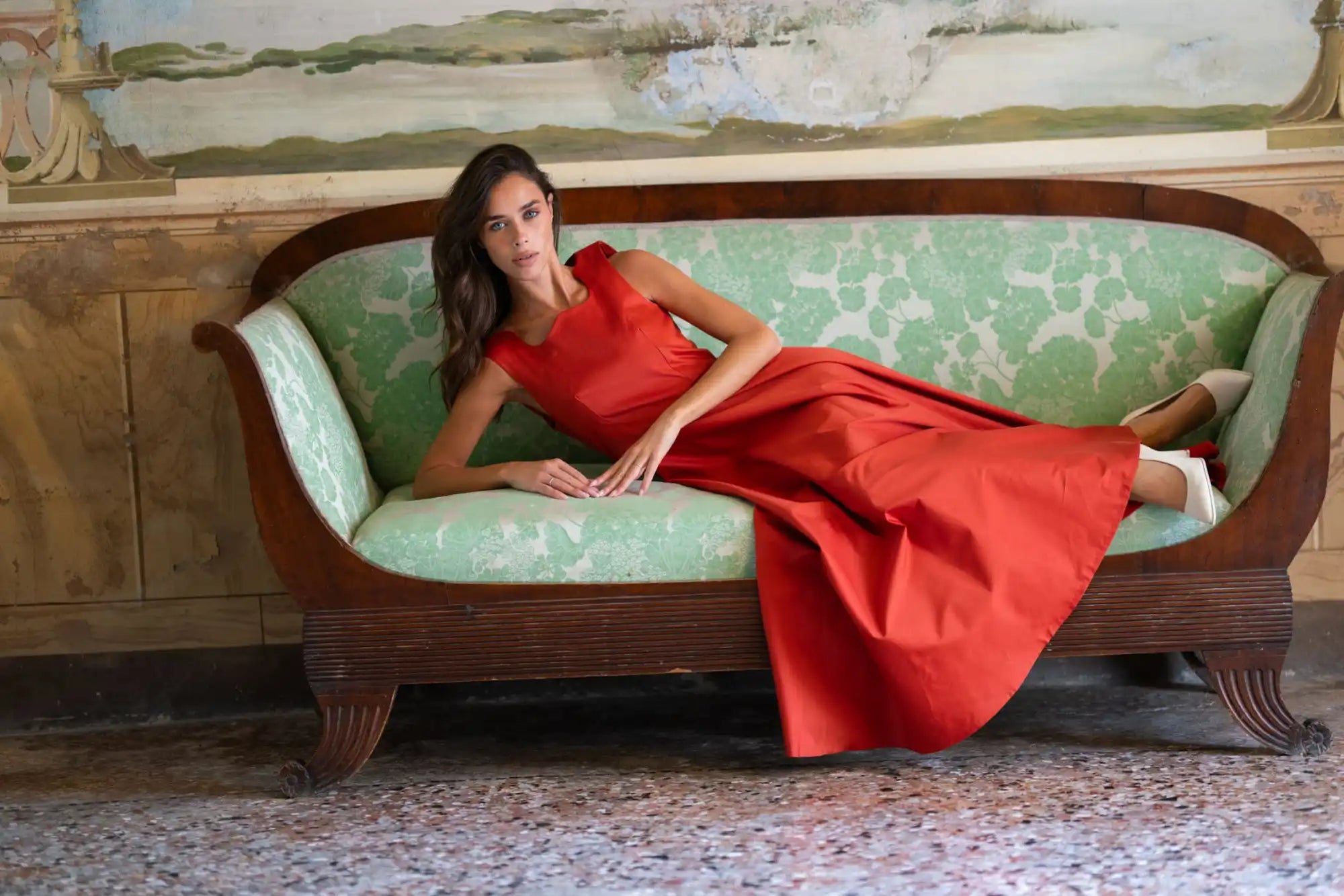 Woman in a flowing red dress lying on a green patterned sofa.